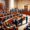 A grand courtroom with numerous people gathered. Judges are seated at a large, curved bench, while others listen intently. Lawyers stand presenting their case. The room is ornate with large columns, drapes, and a flag. Shadows and light highlight the scene.