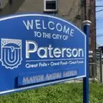 Blue sign with white text reading Welcome to the City of Paterson. Slogans below say Great Falls • Great Food • Great Future. The bottom of the sign reads Mayor Andre Sayegh. In the background are buildings and greenery, highlighting efforts to protect students in Paterson schools.