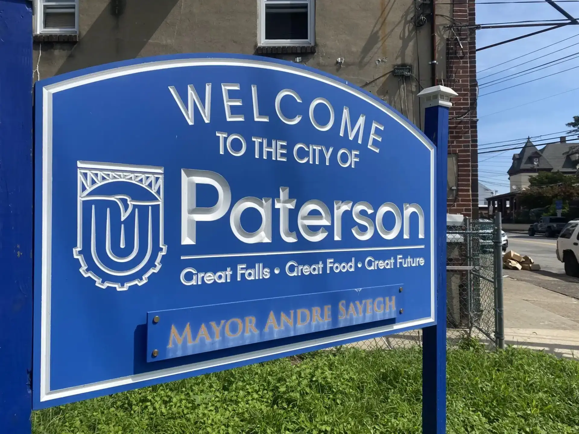 Blue sign with white text reading Welcome to the City of Paterson. Slogans below say Great Falls • Great Food • Great Future. The bottom of the sign reads Mayor Andre Sayegh. In the background are buildings and greenery, highlighting efforts to protect students in Paterson schools.