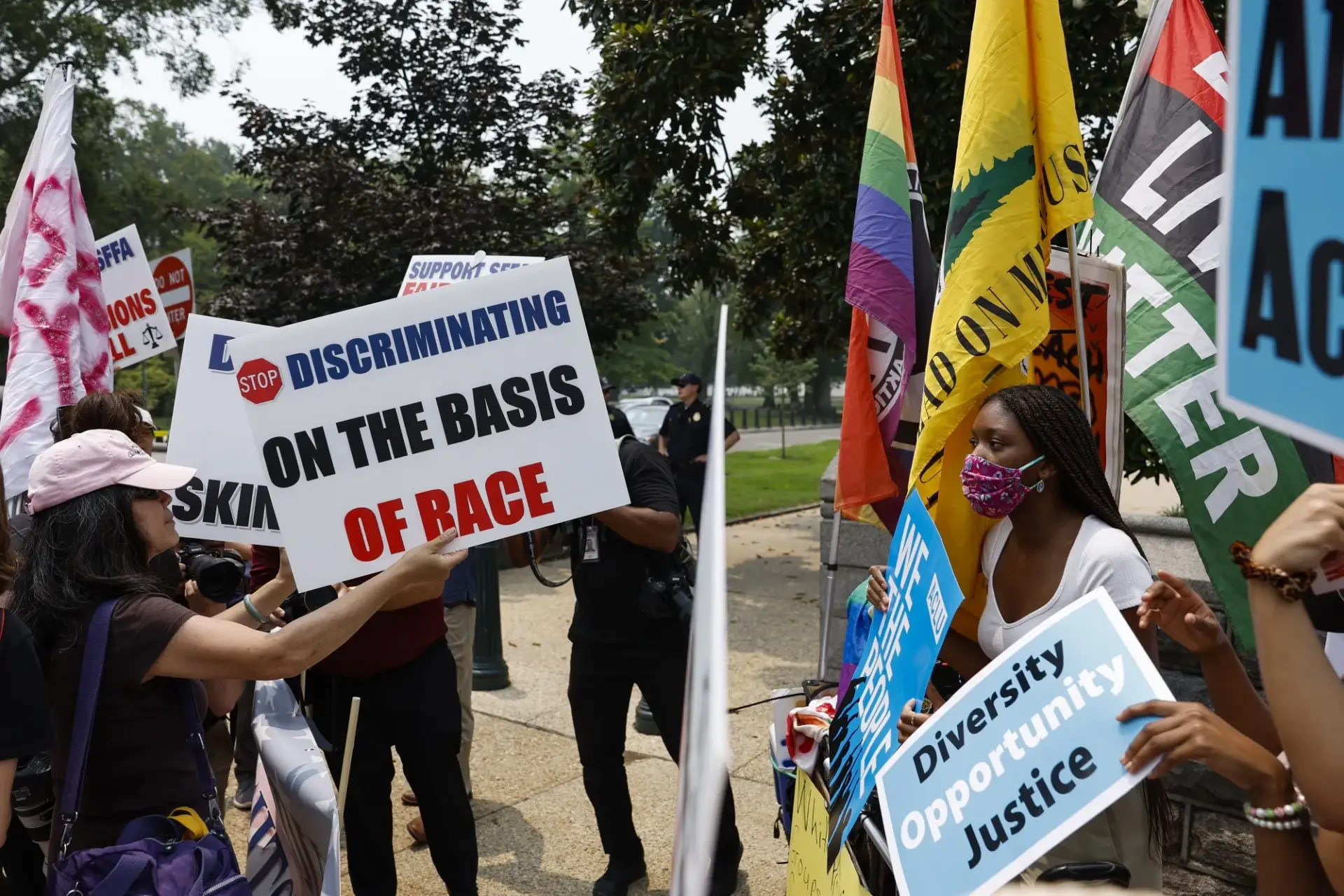A group of protesters engaged in a tense debate over Diversity, Equity, and Inclusion (DEI) policies. On one side, individuals hold signs opposing affirmative action, with slogans like 'Stop Discriminating on the Basis of Race.' On the other side, activists display signs advocating for diversity, opportunity, and justice, alongside flags representing various social movements, including Black Lives Matter and LGBTQ+ rights. The scene captures the deep political and ideological divide over DEI initiatives in the United States